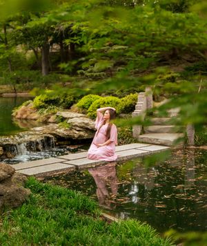 Pregnant woman in pink dress sitting by a serene garden pond with waterfall and lush greenery.