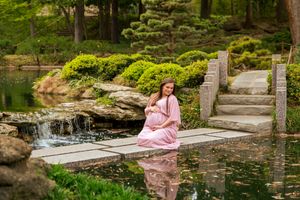 Pregnant woman in pink dress sitting on stone bridge over pond in Japanese garden with waterfall.