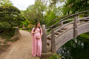 Pregnant woman in pink gown poses by wooden bridge in lush garden during maternity photoshoot.
