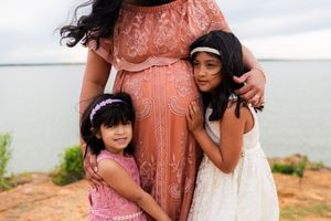 Two young girls in dresses hug a pregnant woman in a pink lace gown outdoors near water.