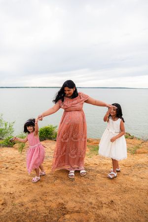 A pregnant woman in a pink dress holds hands with two young girls in dresses near a scenic waterfront.