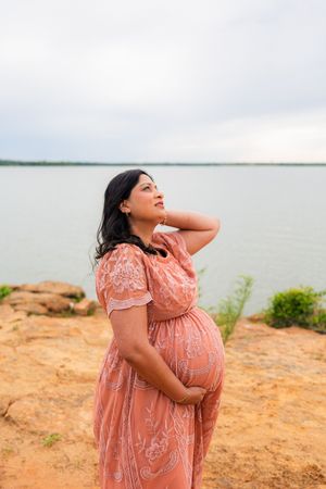 Pregnant woman in pink lace dress cradling belly by lakeside, posing outdoors on rocky shore under cloudy sky.