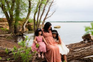 A pregnant mother in a pink dress sits on a log by a lake with her two young daughters in a serene outdoor setting.