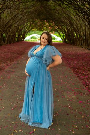 Expectant figure in flowing light blue gown poses beneath natural archway covered with blooming red flowers.