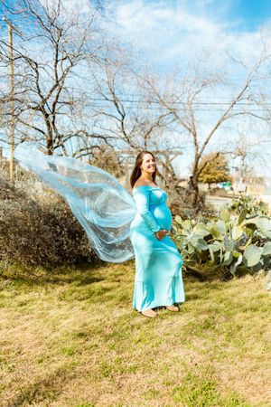 A person in a flowing turquoise dress poses gracefully on a grassy hillside on a sunny spring day.