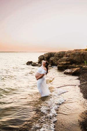 Pregnant woman in white dress standing in ocean waves at sunset near rocky shoreline during maternity photo session.