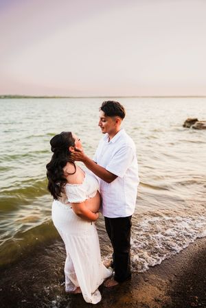 Couple sharing a tender moment on a beach at sunset, woman in white maternity dress, man in white shirt touching her face.