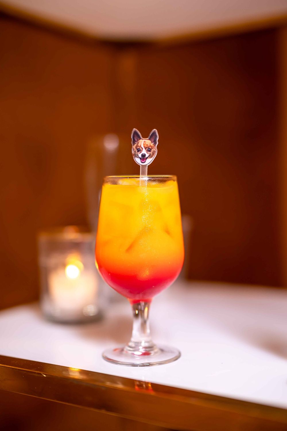 Tropical sunset cocktail in a stemmed glass with a decorative cat-shaped drink stirrer and candle in background.