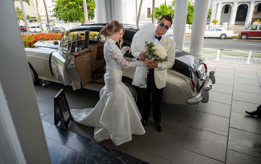 Two people in formal attire stand next to a vintage luxury car under a covered entrance.