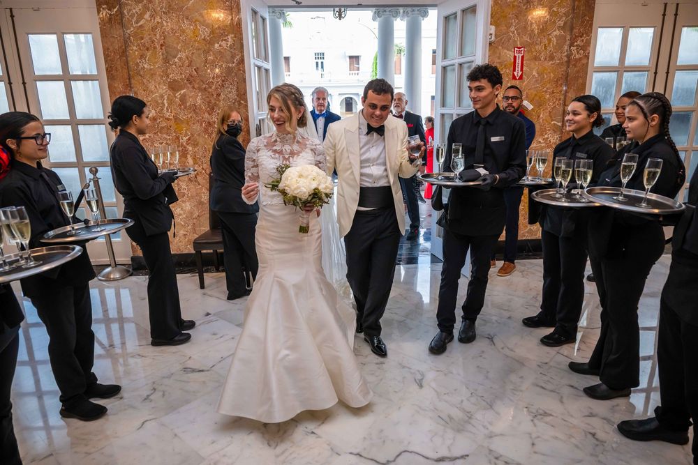 Wait staff in black uniforms serve glasses to a couple at an elegant wedding reception in a stone-walled venue.