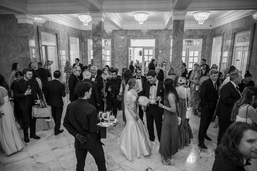 Black and white photo of an elegant wedding reception in a grand marble hall with guests mingling and celebrating.