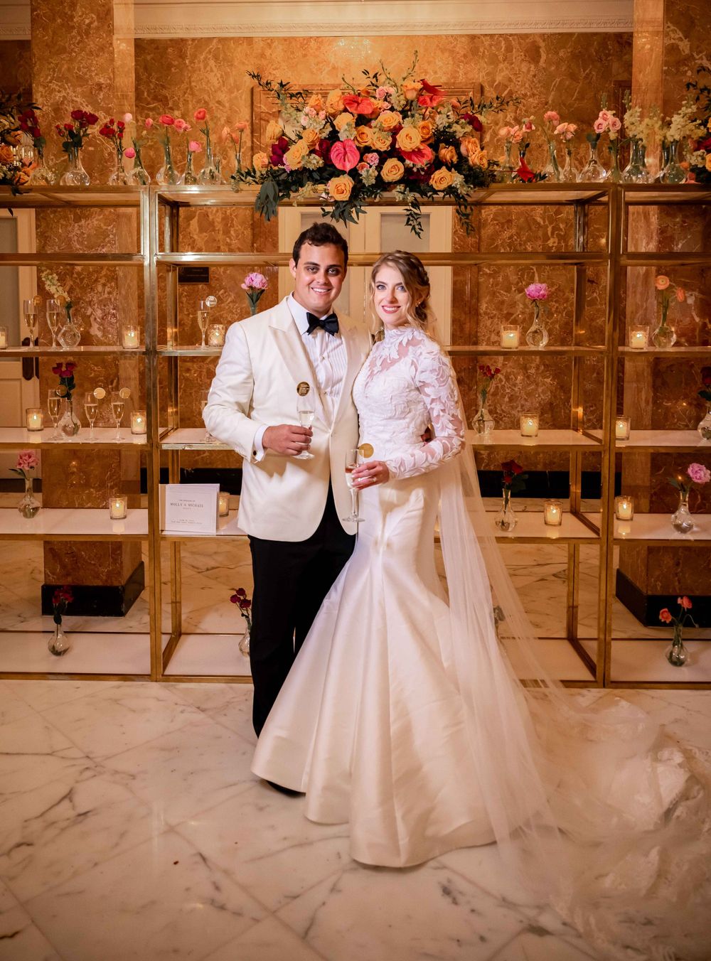 A couple poses in wedding attire in front of a pink floral wall display at an elegant venue.