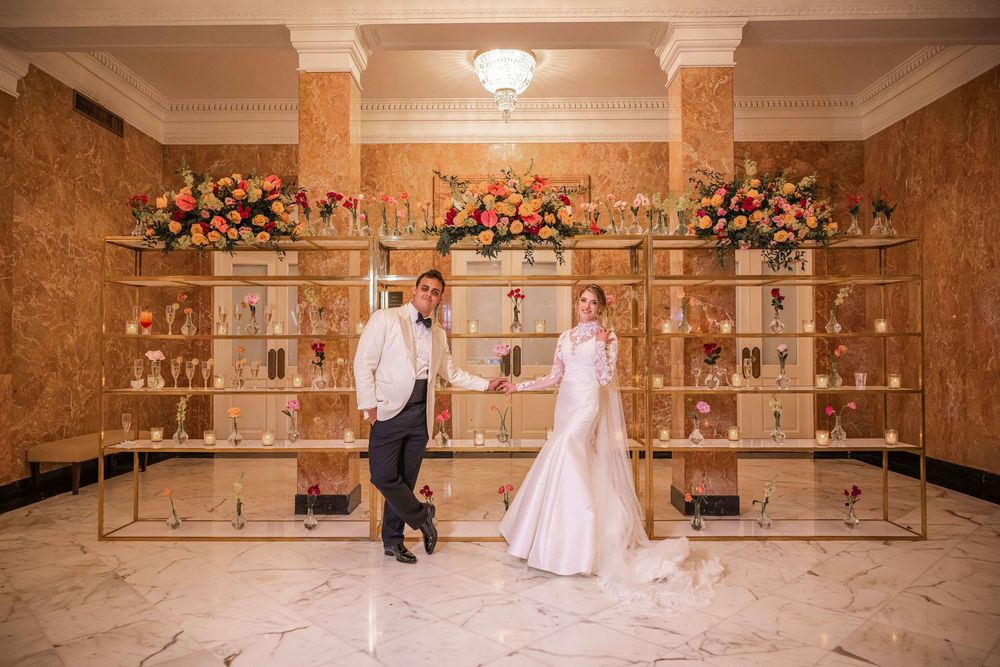 Elegant couple poses in front of ornate display shelves decorated with floral arrangements in luxurious marble venue.