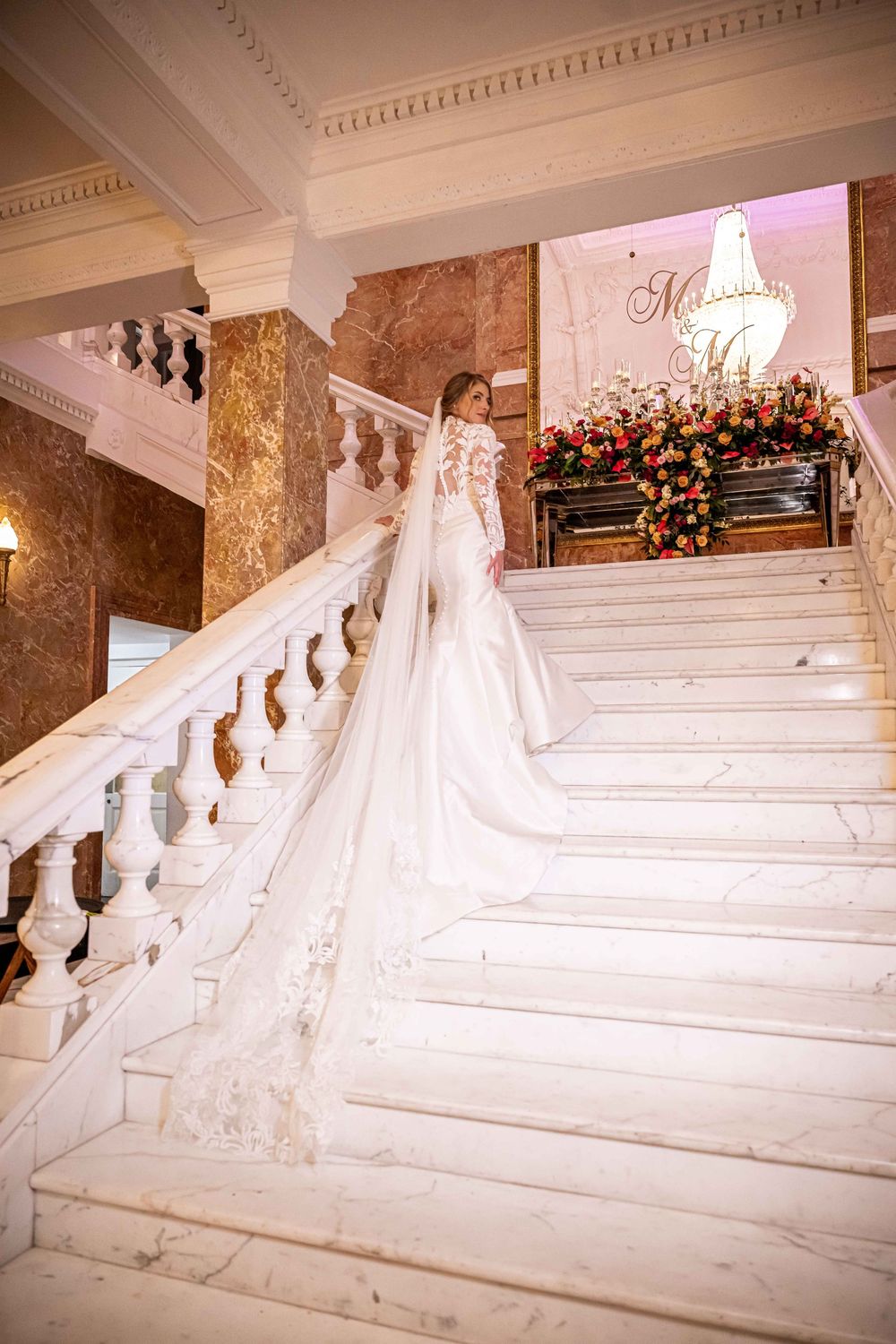 Elegant bridal figure ascending grand white marble staircase with ornate balustrade and floral arrangements in luxurious venue.