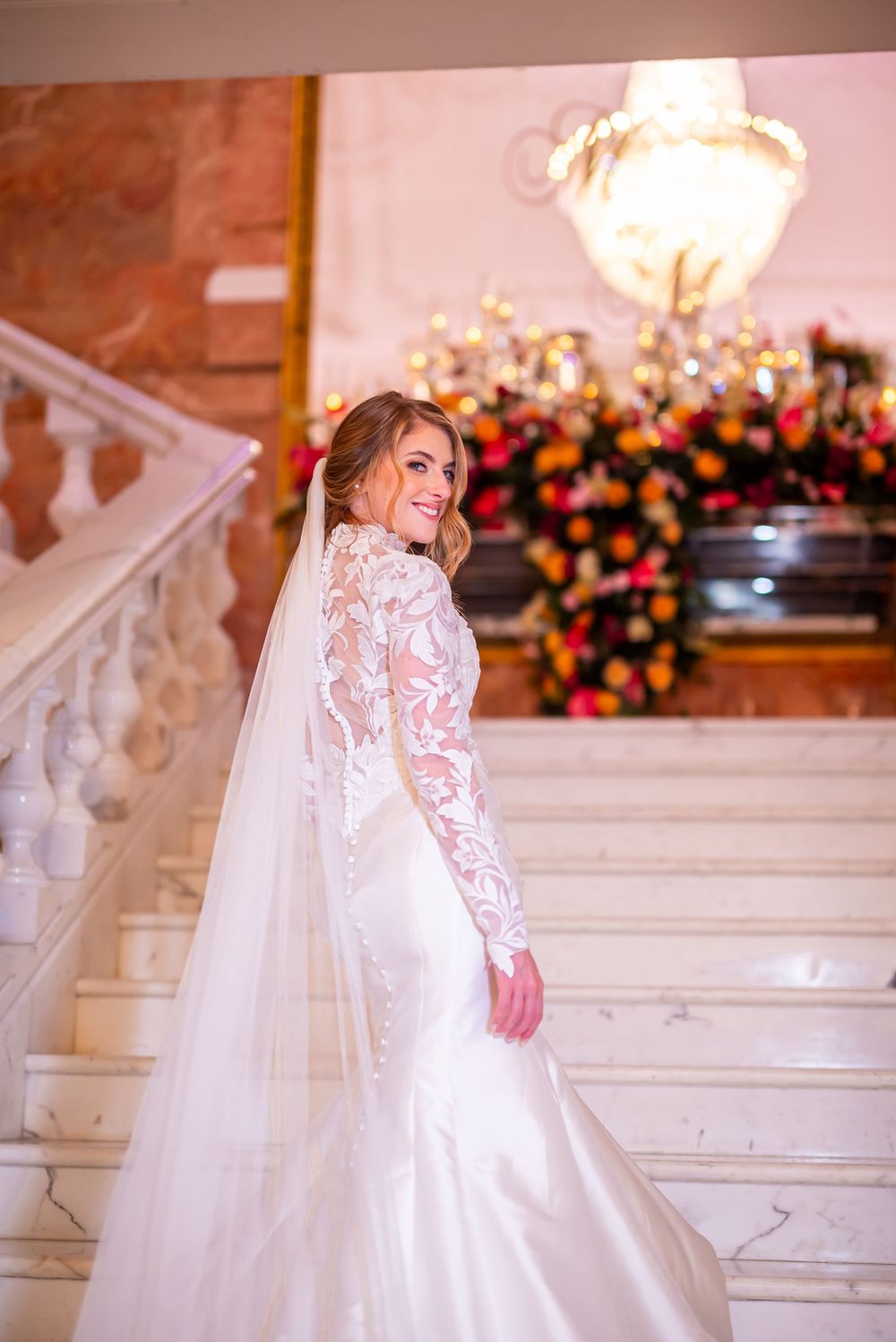 An elegant wedding dress with long lace sleeves and flowing train photographed on marble stairs with floral decor and chandelier.