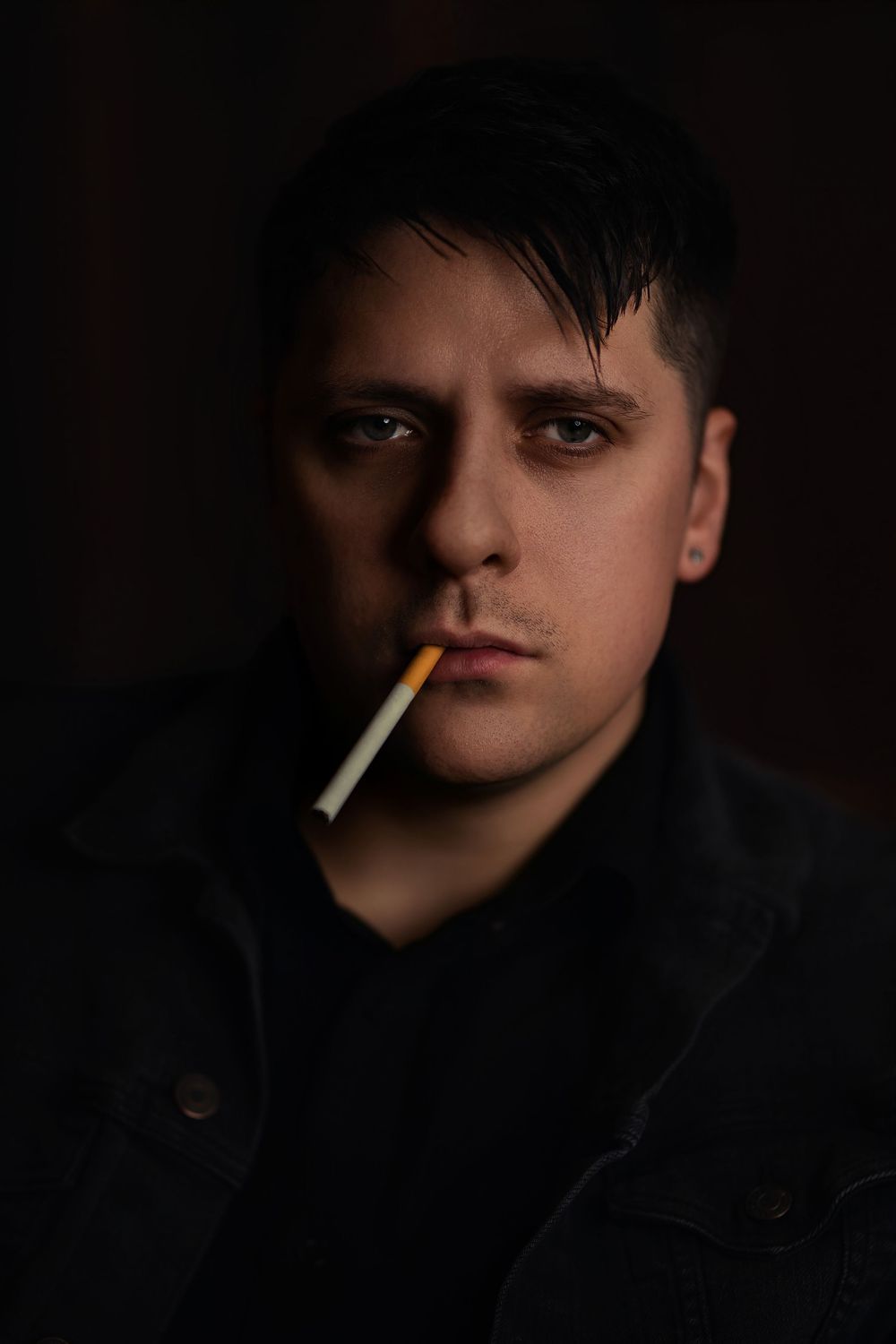 Moody low-key portrait in dark tones showing a cigarette smoker in dramatic lighting against a black background.