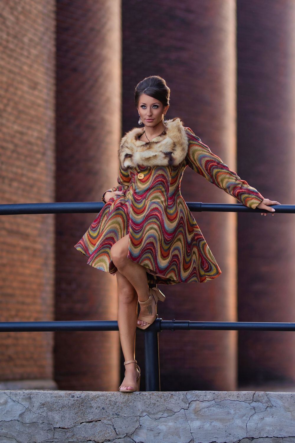 Elegant dancer in wavy patterned dress and fur collar poses gracefully on railing near brick columns.