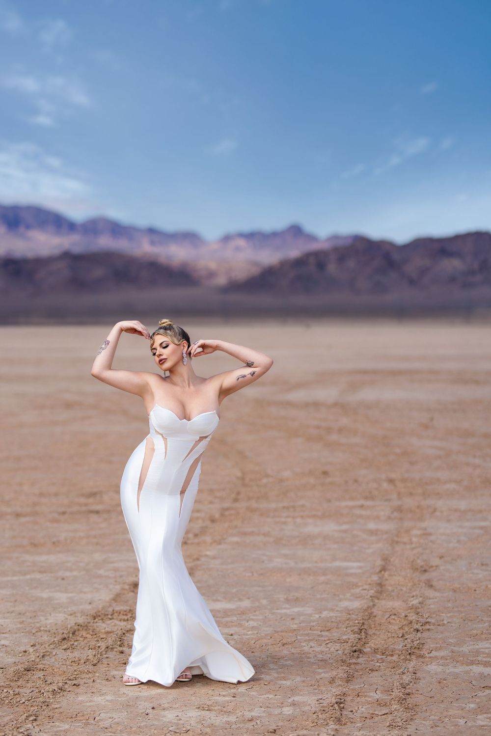 Figure in flowing white gown strikes graceful pose against desert landscape with mountains in background.