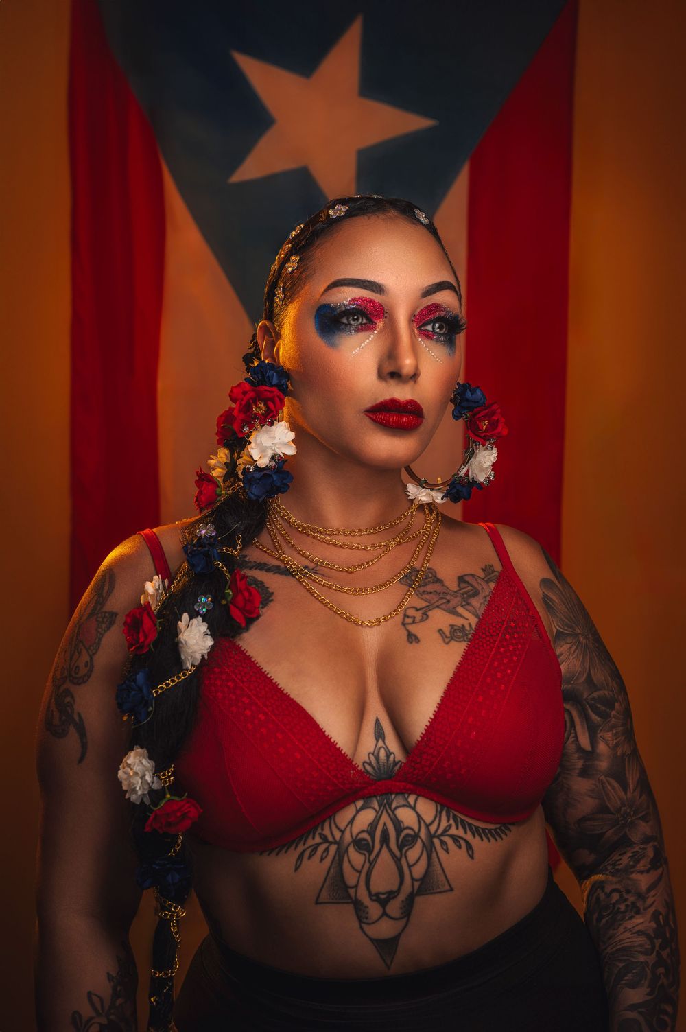 Artistic portrait with Puerto Rican flag backdrop featuring vibrant makeup, red attire, floral hair accessories and tattoos.