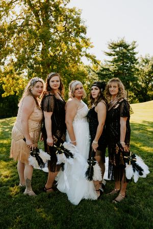 Wedding party wearing black and beige dresses pose with feather boas on grassy hill.