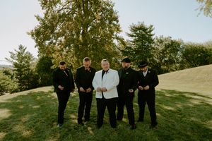 Groomsmen in black suits pose on rolling green hill during sunset.