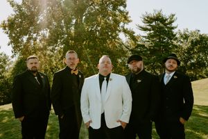 Wedding party members in formal attire gather on grassy slope during golden hour.