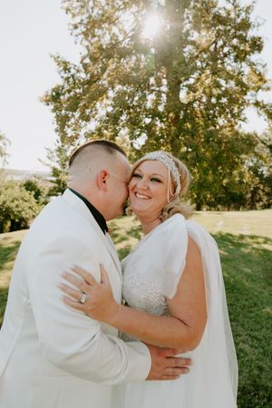 A romantic wedding couple shares an intimate moment under sunlit trees in an outdoor setting.