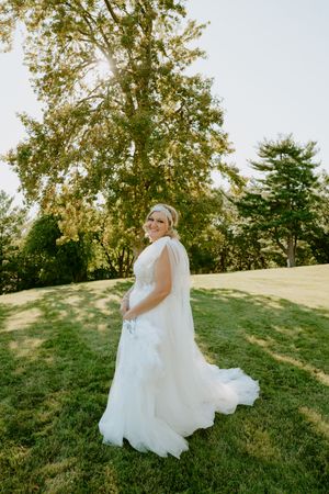 A bride in a flowing white gown poses gracefully on a sun-drenched hillside.