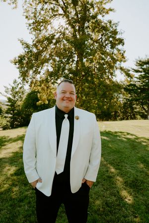 A person in a white tuxedo jacket and black pants stands on a grassy slope with trees in the background.