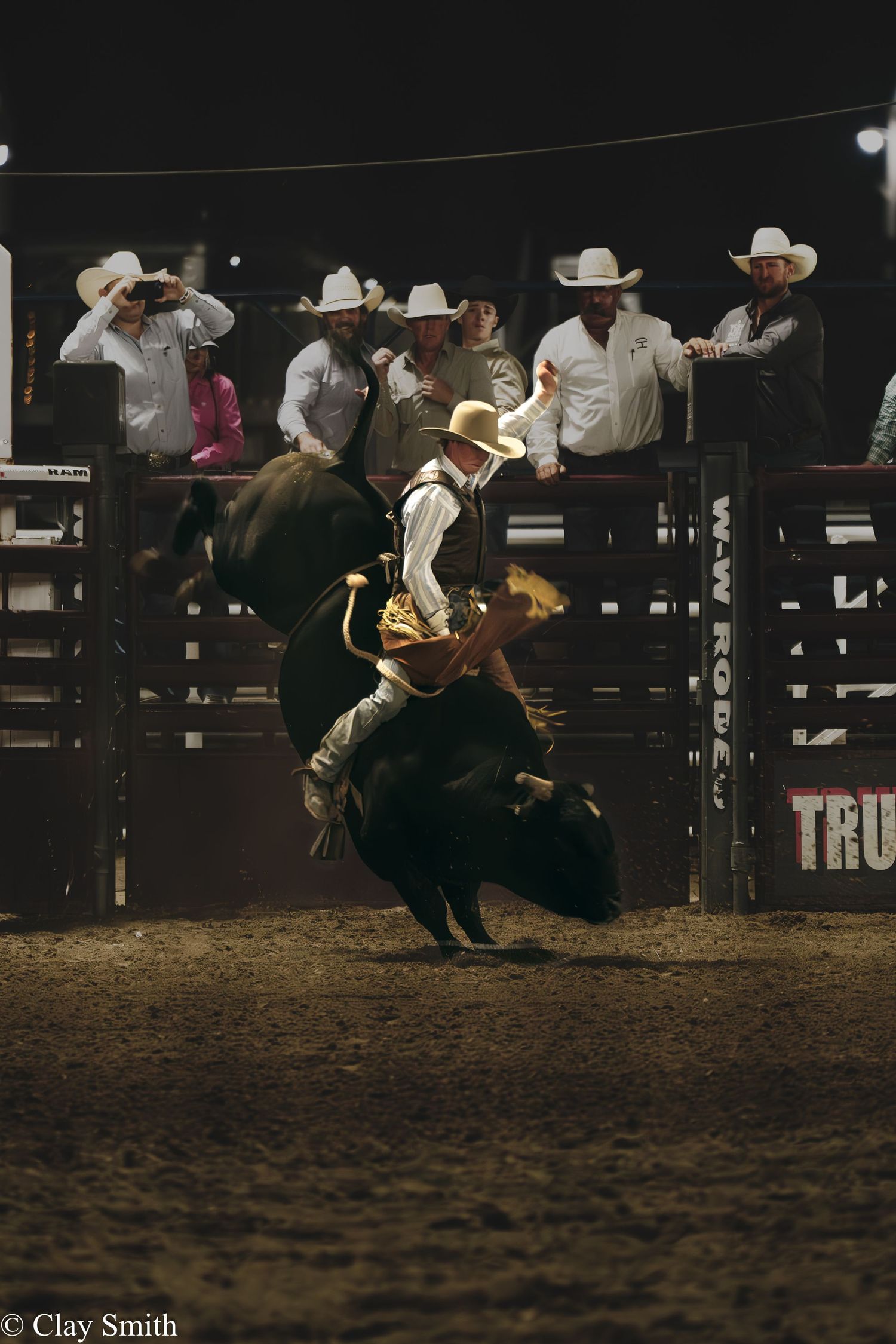 A bull rider competes in a rodeo arena at night while cowboys watch from behind the chutes.