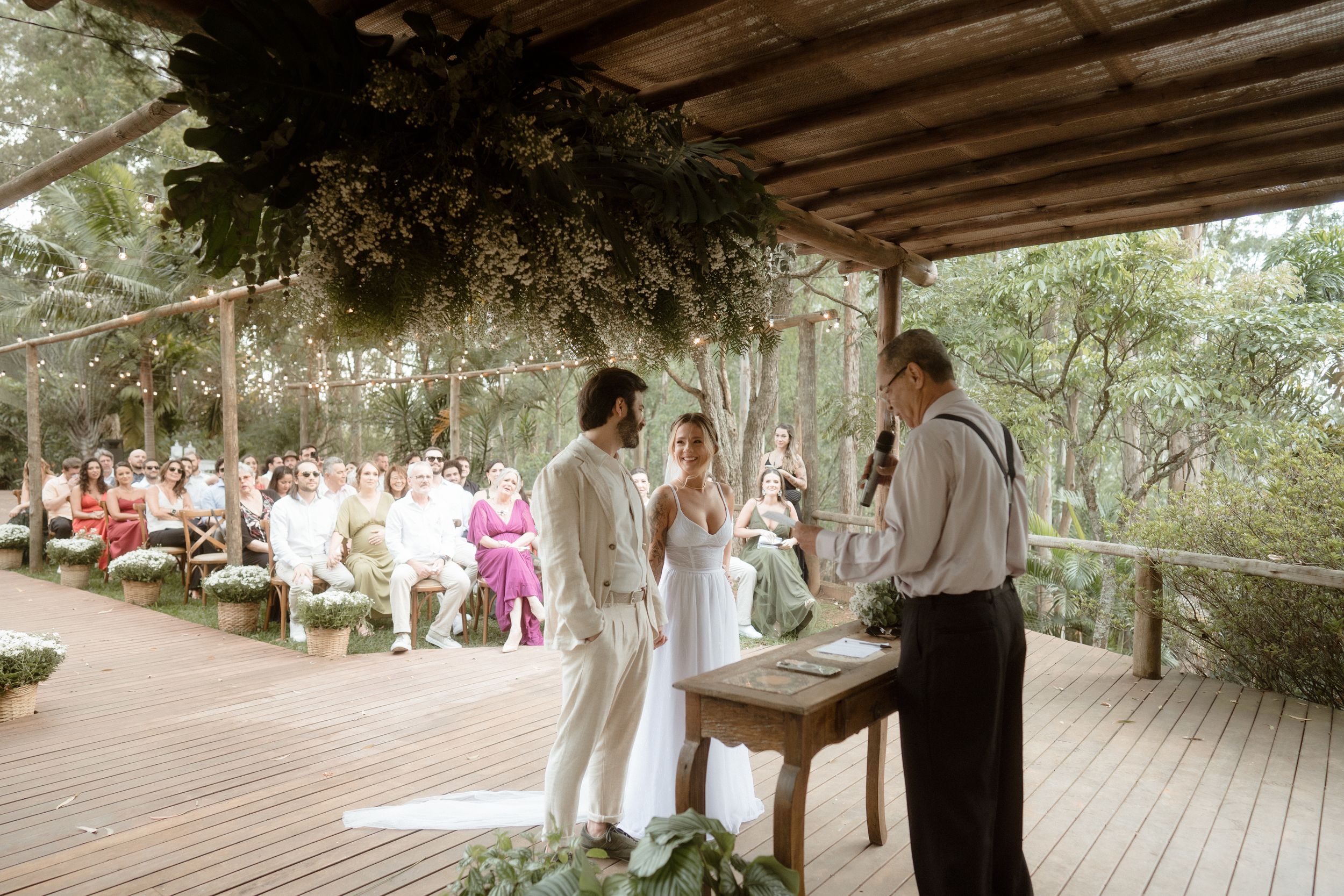 An outdoor wedding ceremony takes place on a wooden deck under a rustic pergola covered in greenery.
