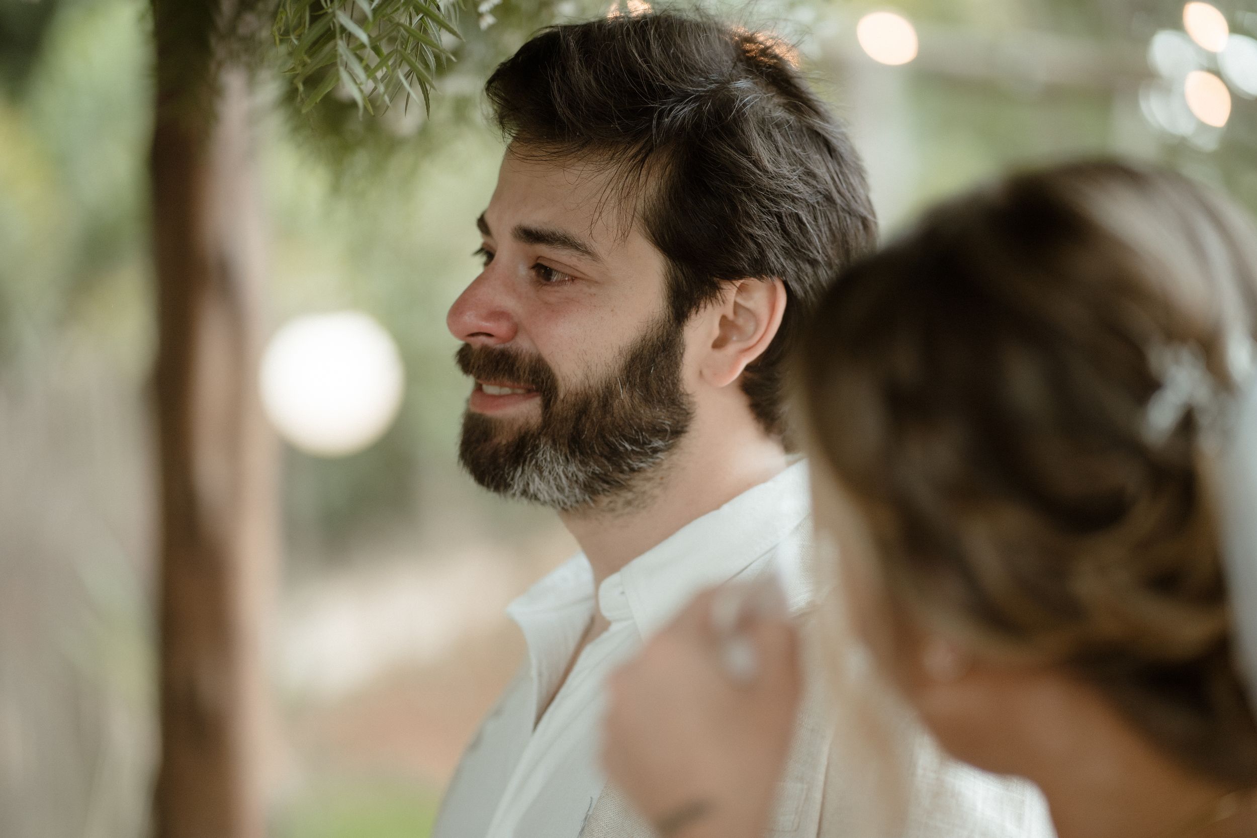 A bearded groom in white attire smiles warmly during an outdoor wedding ceremony.