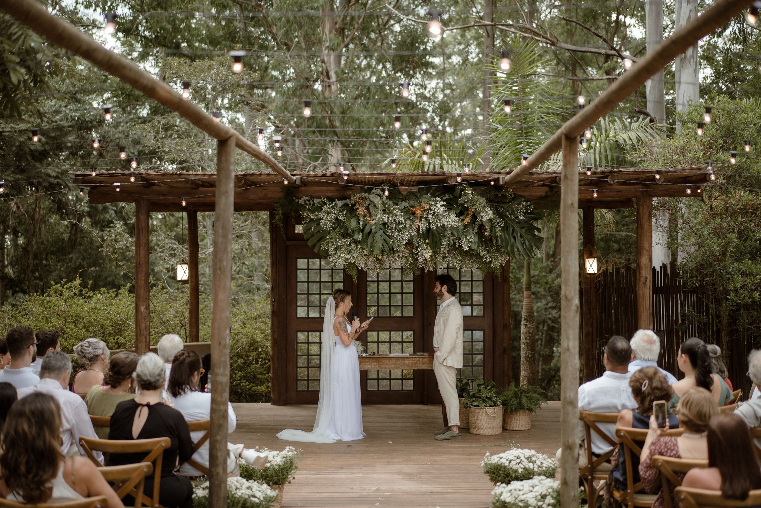 A wedding ceremony unfolds on a wooden deck surrounded by trees with guests seated on wooden chairs.