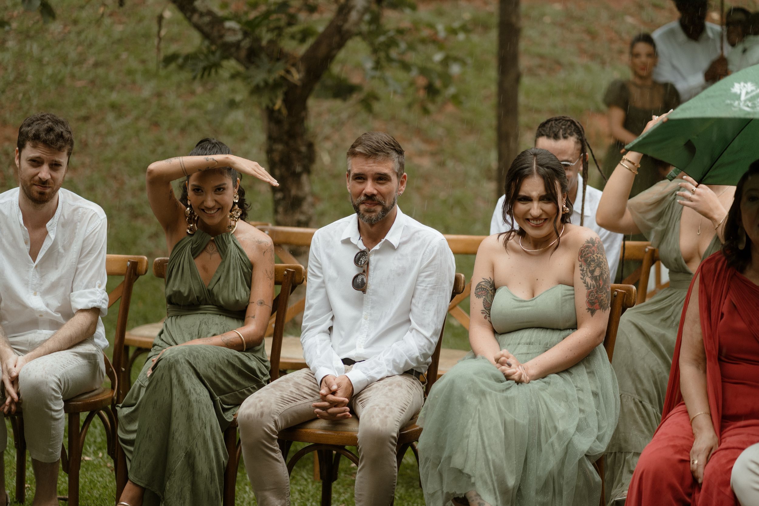 Wedding guests in casual attire sit on wooden chairs during an outdoor garden ceremony.