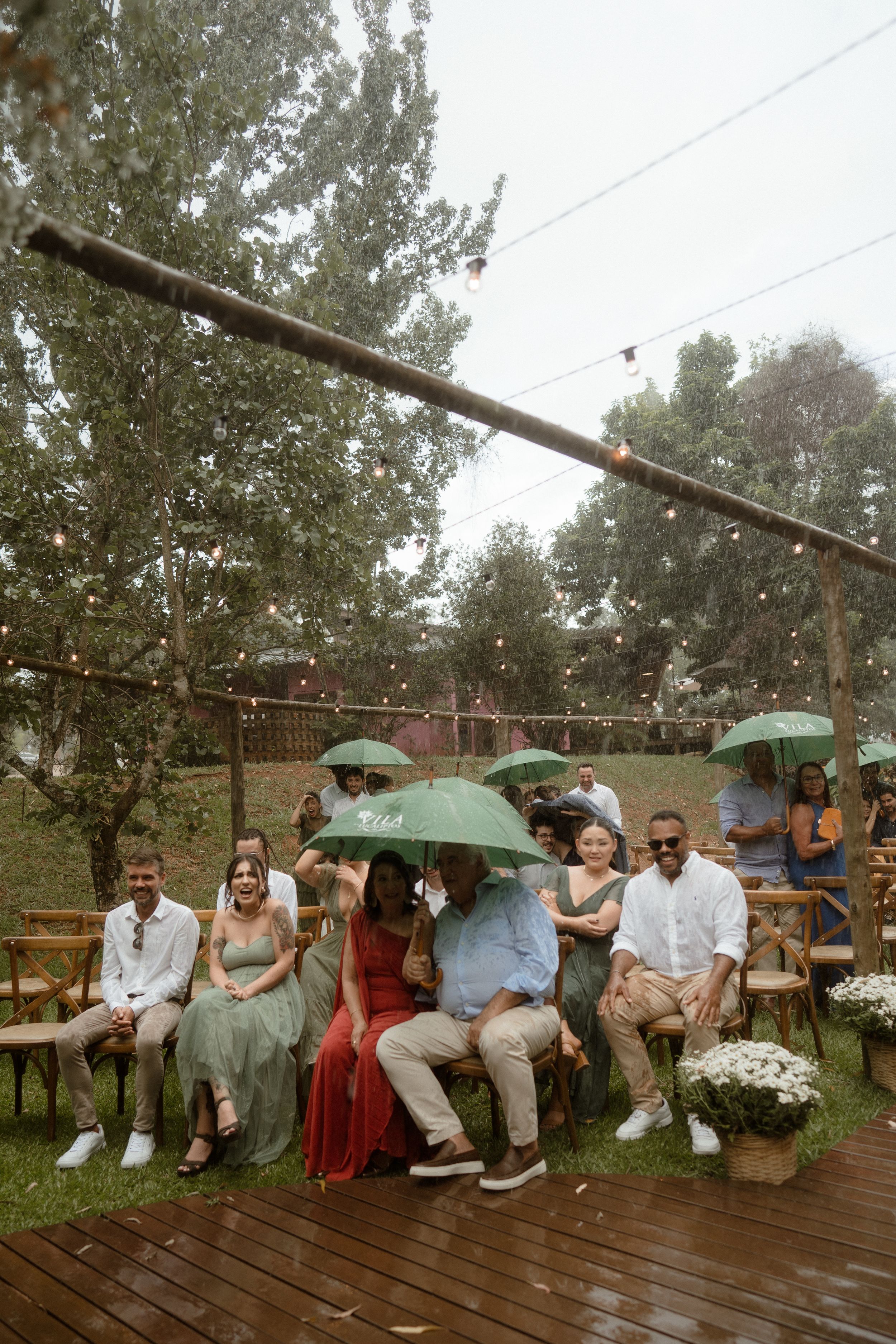 Wedding guests shield themselves with green umbrellas during a rainy outdoor ceremony.