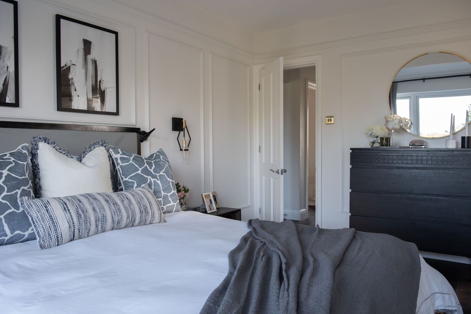 Light and airy panoramic view of a modern bedroom with white walls, plush bedding, and dark furniture accents.