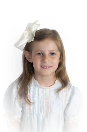 Child in white dress and bow smiles sweetly against white background.