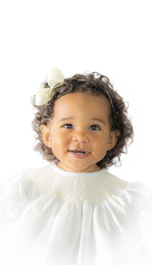 Child in white smocked dress and bow smiles brightly against white backdrop.