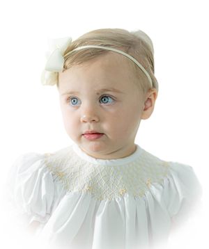 A baby in a white smocked dress and headband poses for a portrait against a light background.