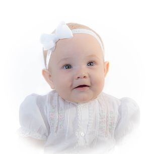 A baby wearing a white embroidered dress and bow headband smiles at the camera.