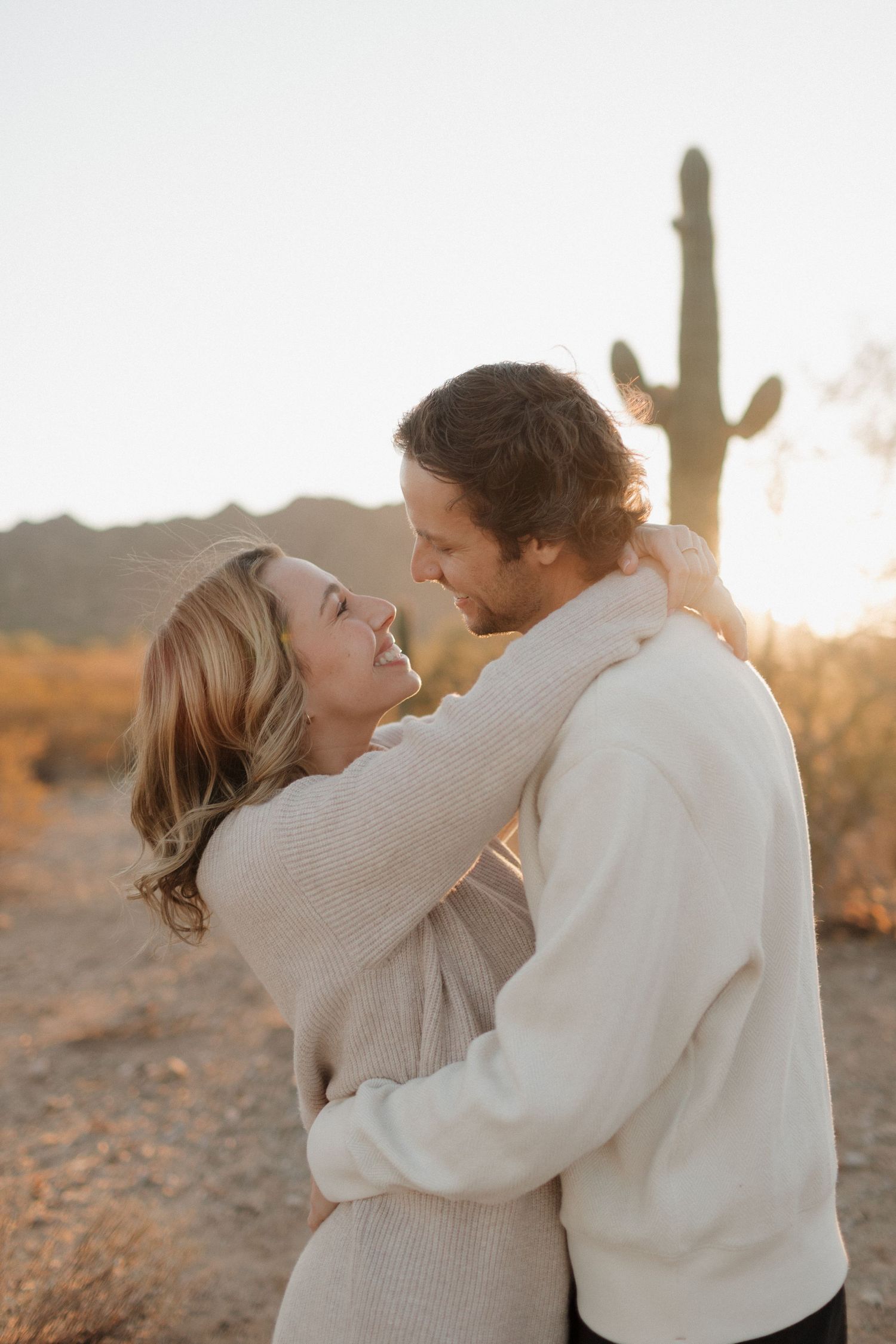 Couple embracing in warm sweaters against desert sunset with saguaro cactus in background.