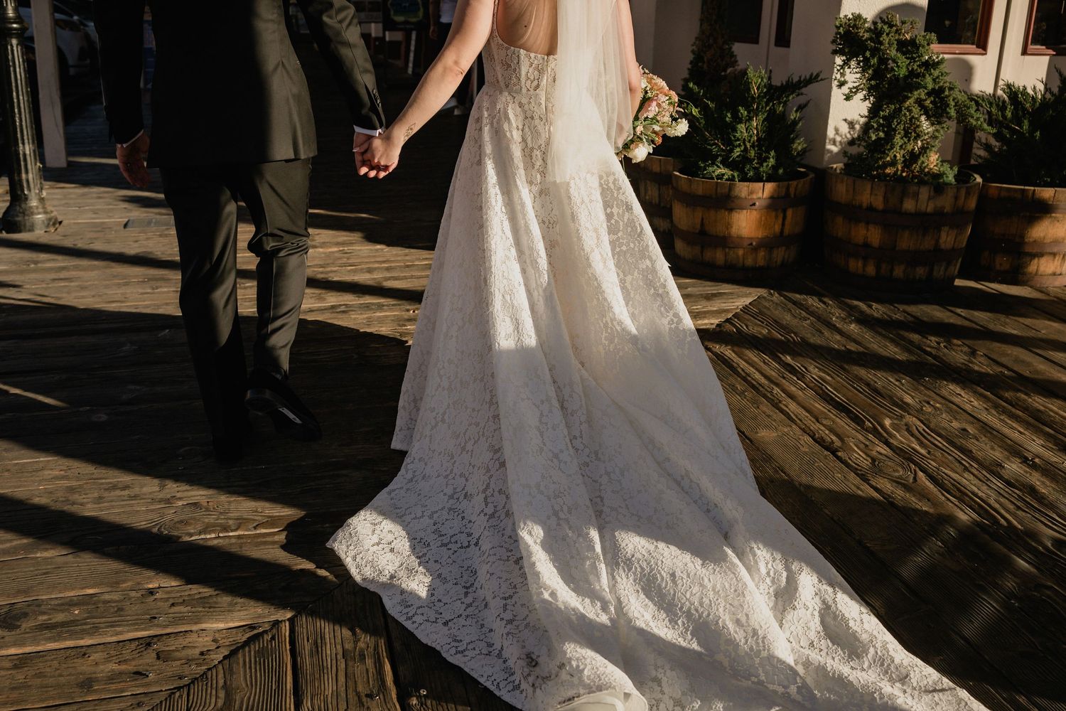 Long white lace wedding dress train trailing on wooden deck in warm sunlight beside rustic barrel planters in Old Sacramento.