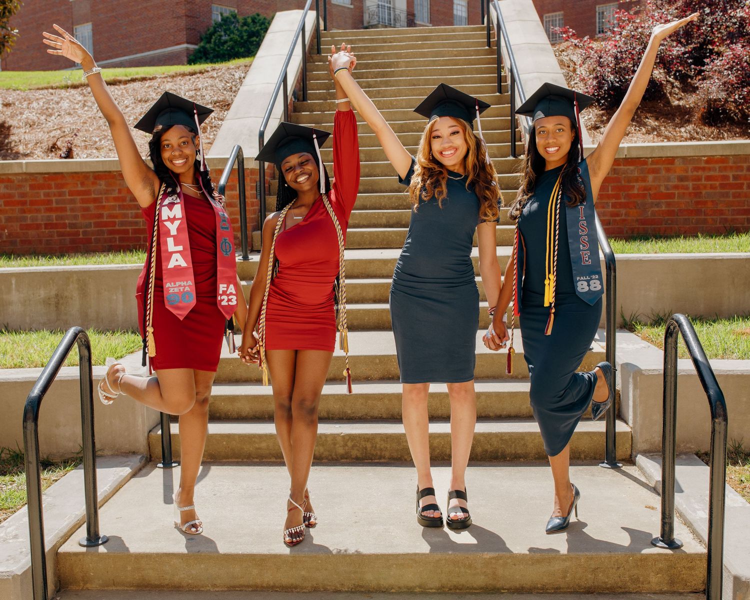 Four graduates in caps and gowns celebrate on campus steps with raised arms.