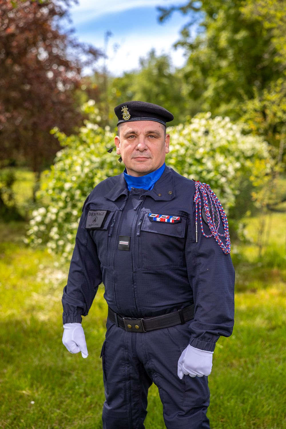 Person in navy blue uniform with decorative patches and white gloves standing in a garden setting.