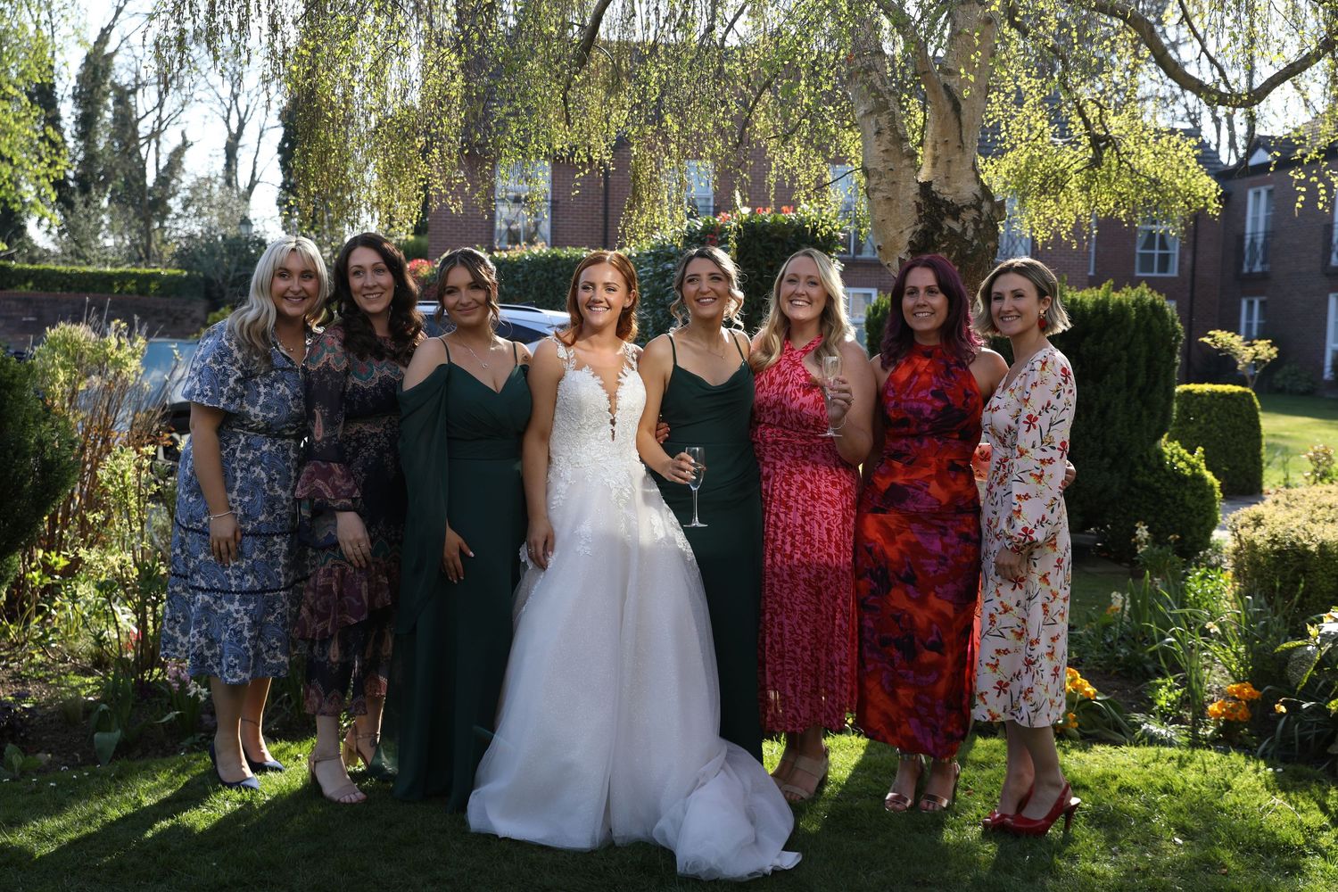 Group of women in colorful dresses pose with bride in garden setting at wedding celebration.