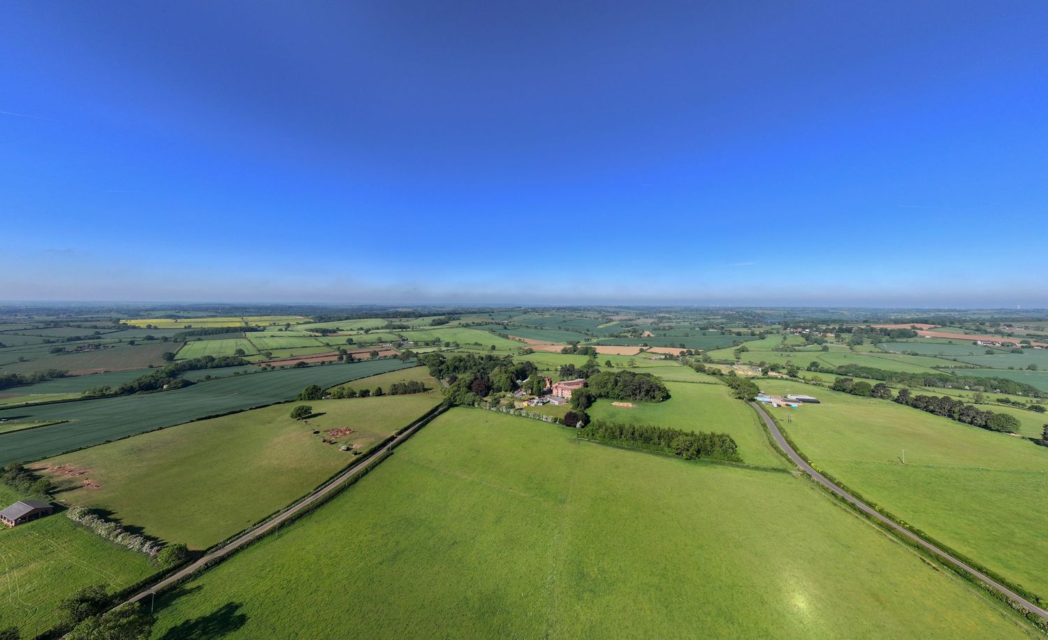 Aerial drone view of verdant green farmland and patchwork fields stretching to the horizon under bright blue summer skies.