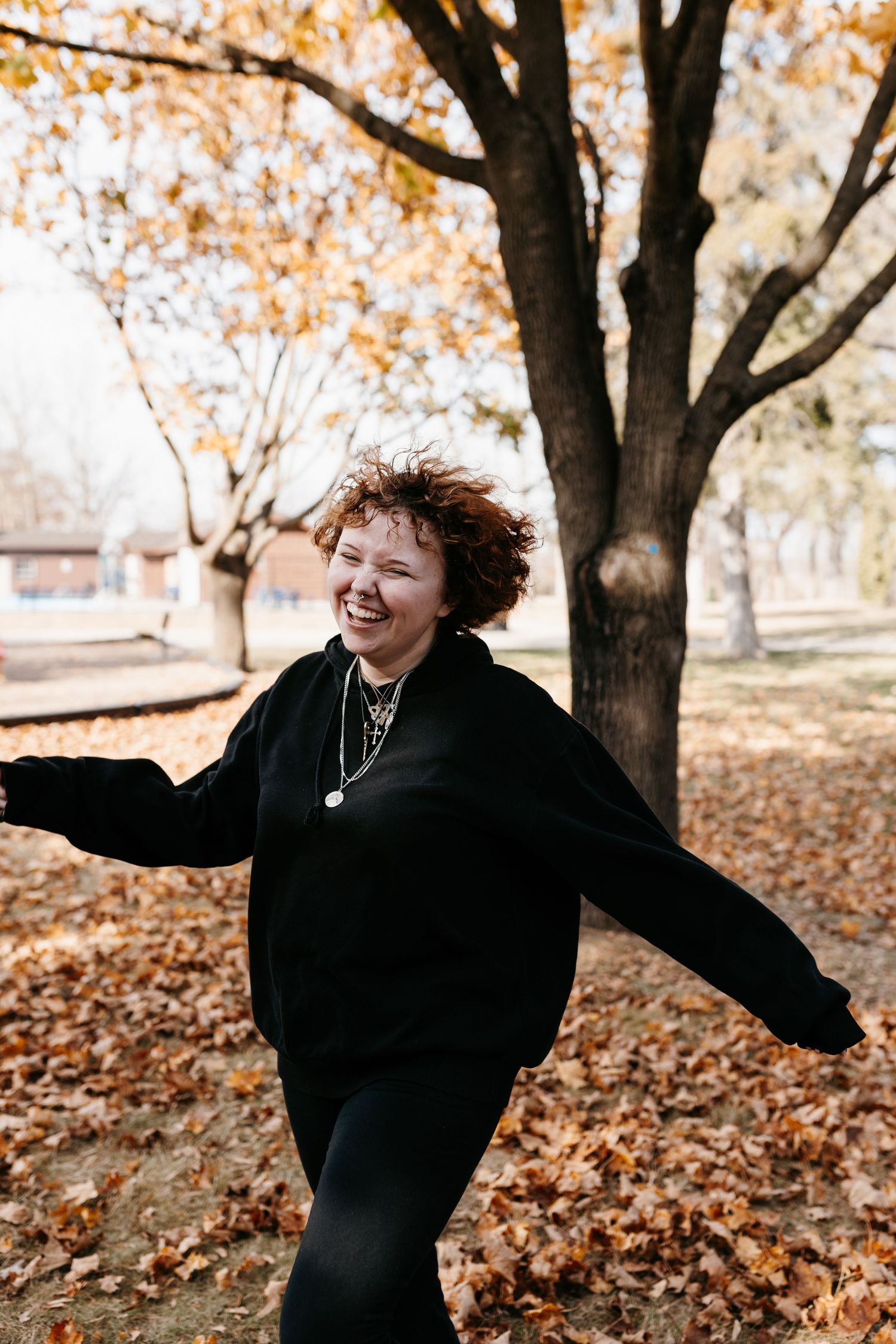 Someone dances playfully in fallen autumn leaves while wearing all black clothing on a crisp fall day.