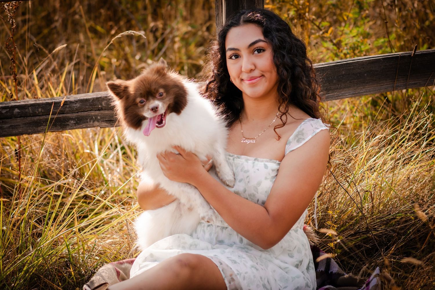 Person in white dress holding fluffy brown and white cat outdoors by wooden fence.