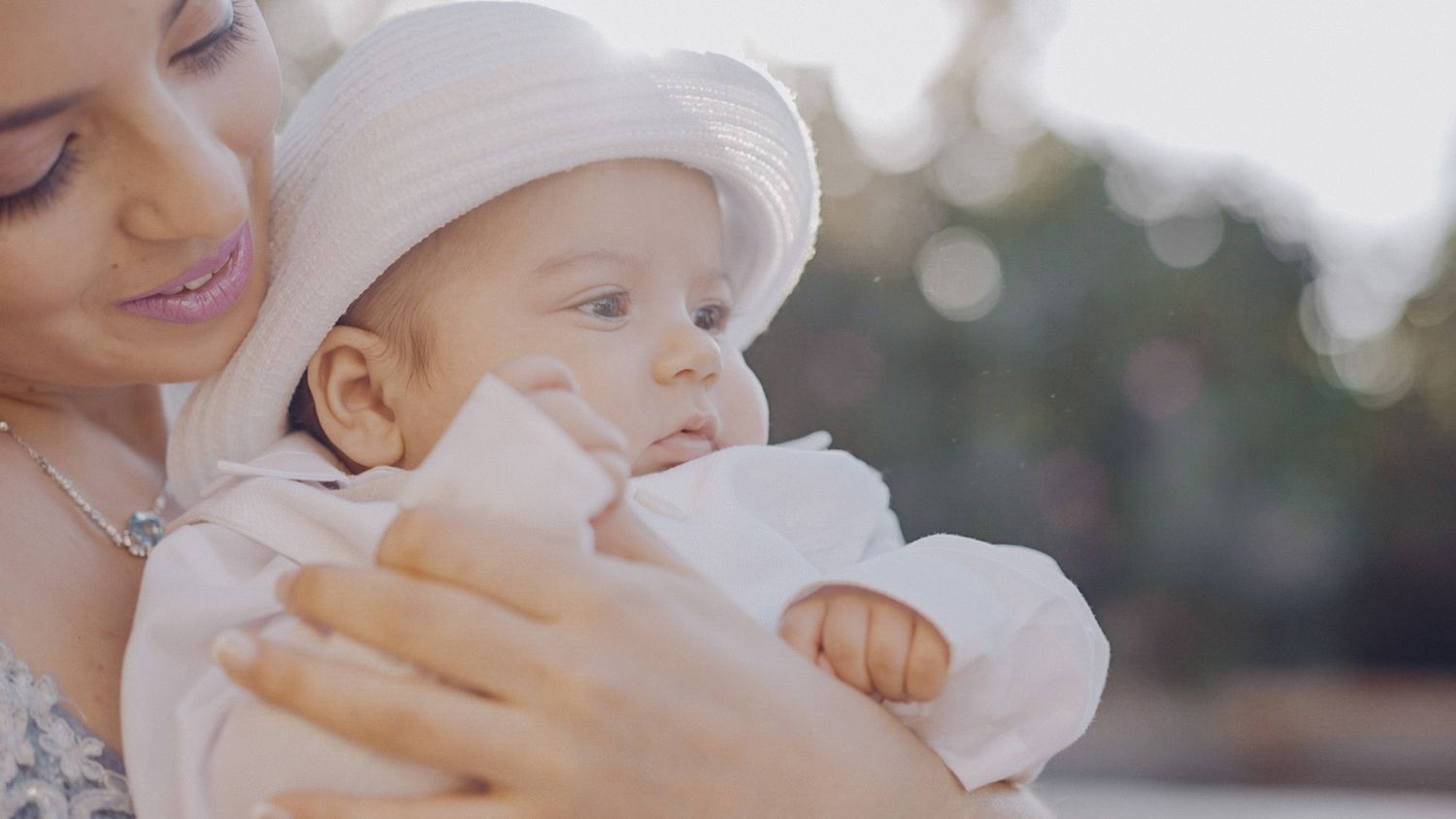 Baby in white clothing is held lovingly in a gentle embrace outdoors with soft sunlight and bokeh effect in background.