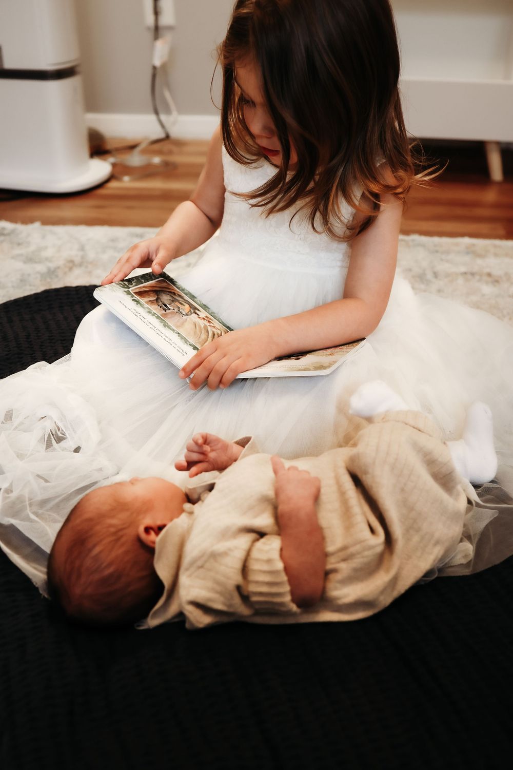 A young reader in white dress sits on a black blanket next to a sleeping baby while looking at a picture book.